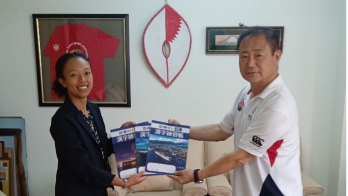 A woman in formal attire hands over Kanji practice notebooks to an older man wearing a sports polo shirt inside a well-lit room. Behind them, a framed red shirt and a decorative shield are displayed on the wall.