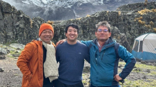 Three hikers posing happily at a snowy base camp on Mount Kenya with dramatic peaks in the background.