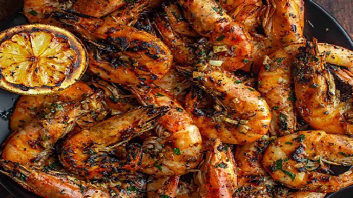 A plate full of grilled prawns seasoned with herbs, served with a grilled lemon slice.