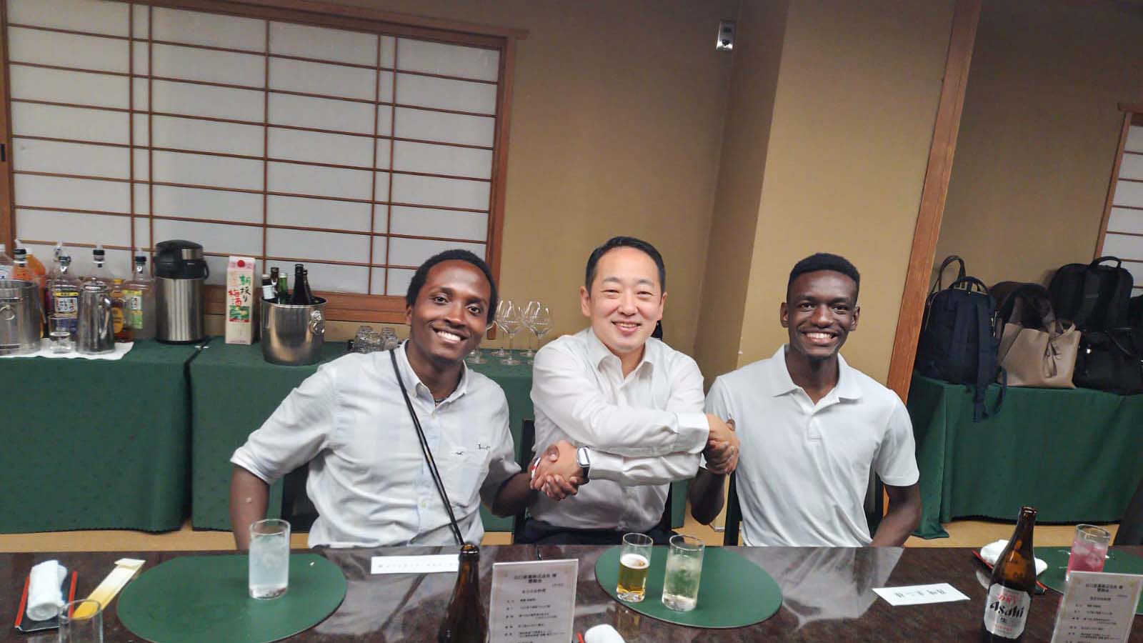 Dinner reception scene with Baraka and Luwate smiling and shaking hands with Yamaguchi Sangyo representatives, fostering cultural exchange.