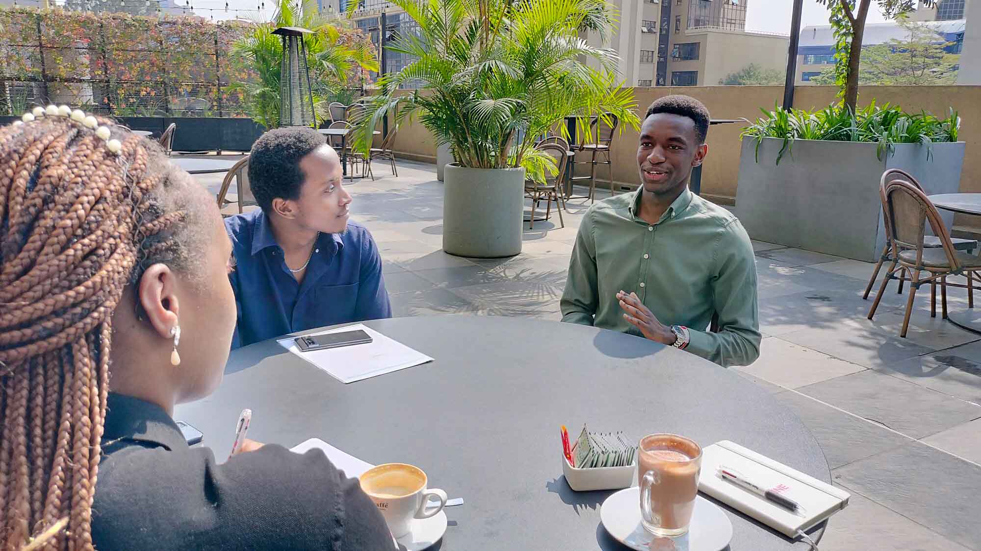 Three Kenyan learners sit outdoors at a café table, discussing their Japanese language journey, with notebooks, coffee, and sunlight around them.