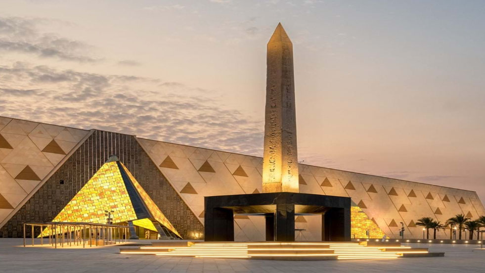 夕暮れ時のギザにある大エジプト博物館の外観。ピラミッドをモチーフにした近代的な建築と、黄金色にライトアップされたオベリスクが印象的。