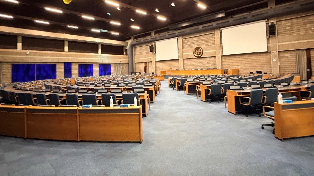 Large conference hall at the United Nations Office in Nairobi, with rows of desks, chairs, presentation screens, and the UN emblem.
