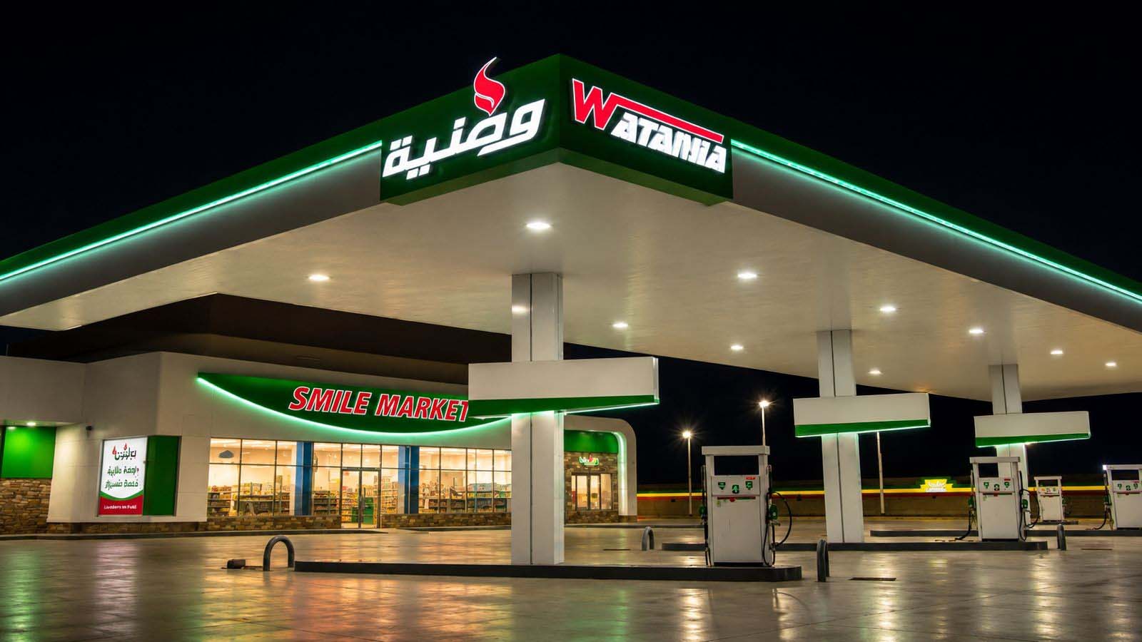 An illuminated Wataniya fuel station and Smile Market convenience store at night.