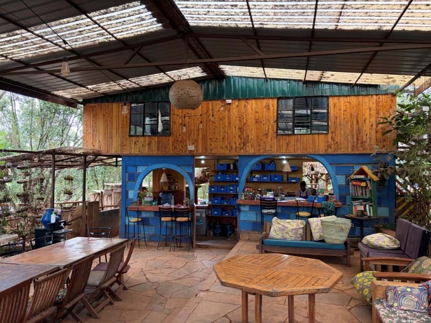 Rustic communal dining and lounge area at Mlango Farm in Kenya, featuring wooden furniture, colorful blue walls, and an open-air design surrounded by greenery.