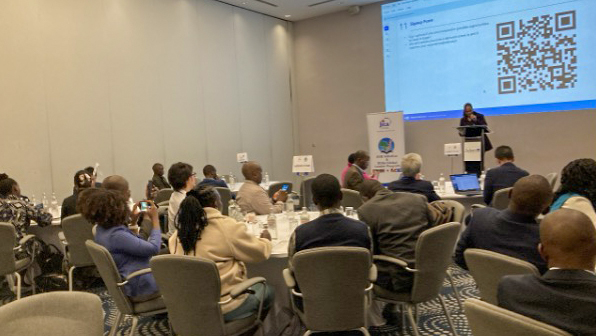 Audience seated during a presentation, with a QR code displayed on the screen and a speaker at the podium.