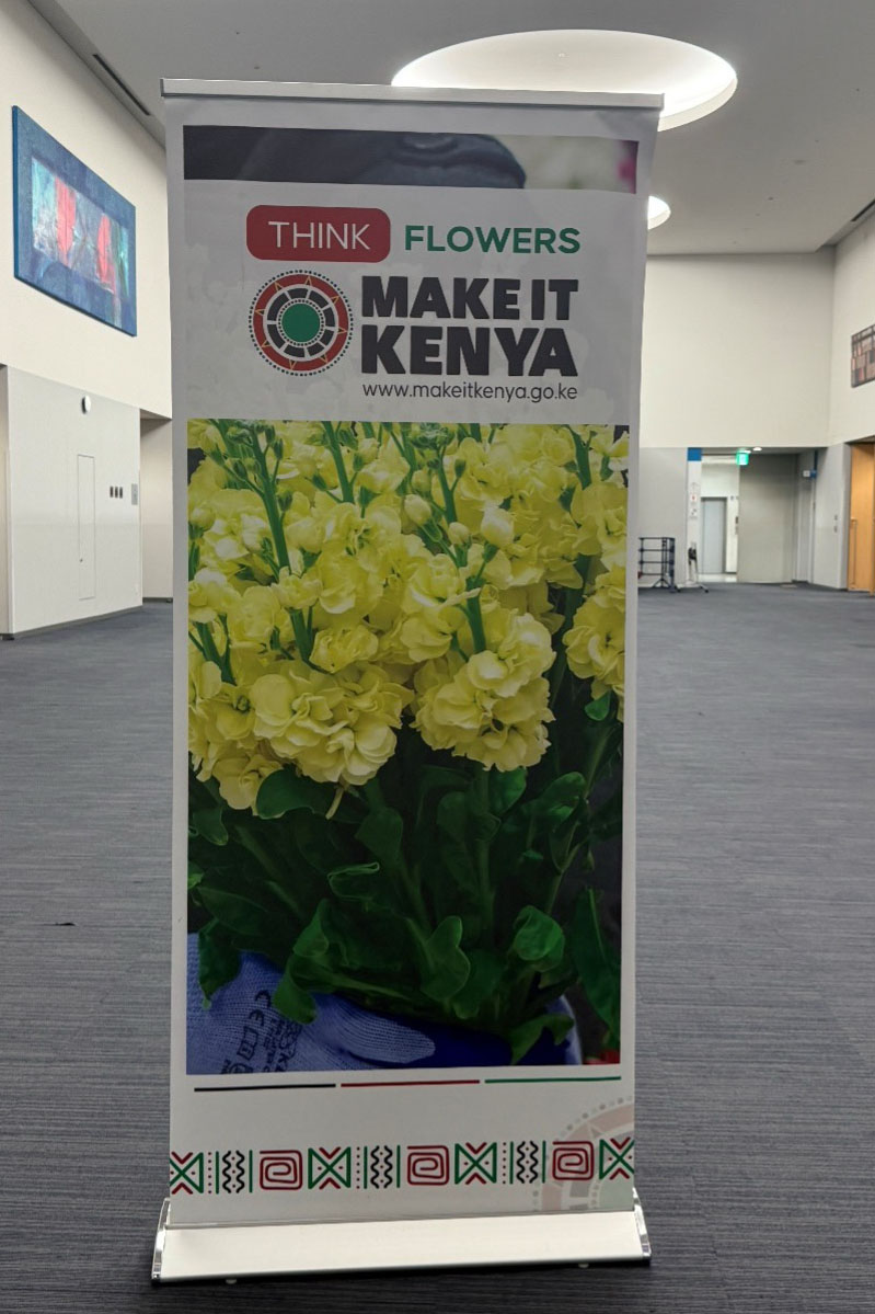 EXPO2025の会場内の廊下に設置された「THINK FLOWERS – MAKE IT KENYA」のプロモーションバナー。黄色い花と公式ウェブサイトの情報が掲載されている。