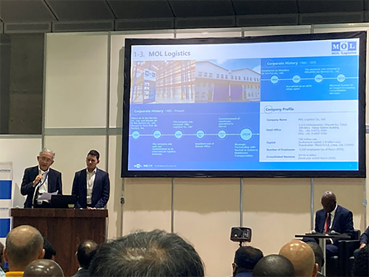 MOLロジスティクスのプレゼンテーション。大画面に会社の沿革と概要が映し出され、登壇者が説明している。