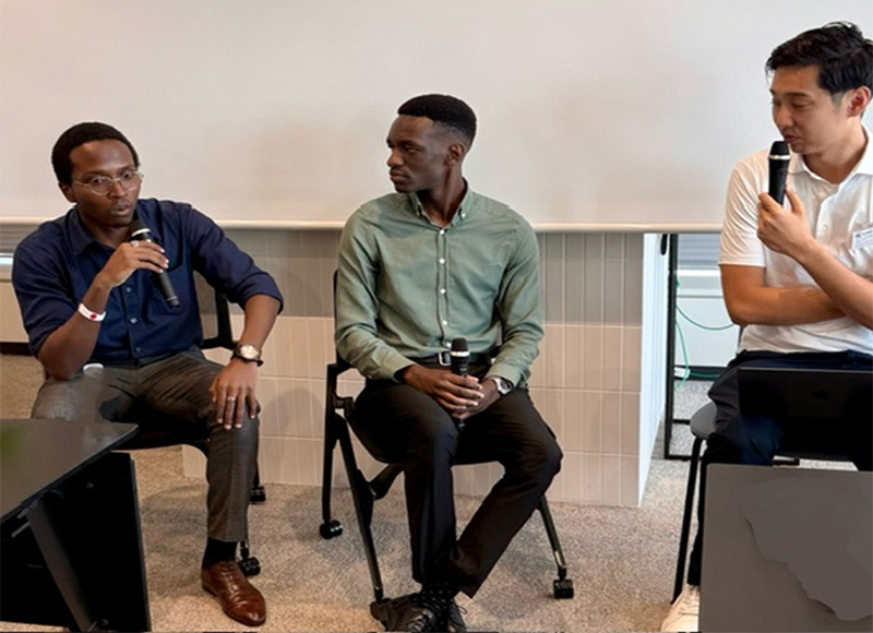 Two Kenyan professionals and a Japanese moderator sit on stage with microphones, participating in a panel discussion.